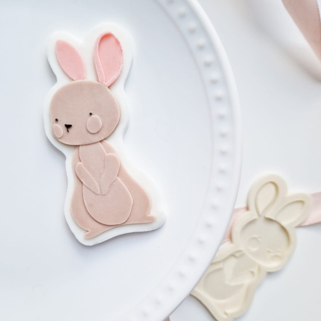 A white ceramic plate with a scalloped edge holds a pastel pink bunny-shaped cookie cutter with large ears and a cute face. A white bunny cutter and pink ribbon are visible in the blurred background.