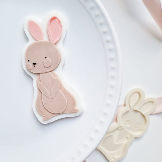 A white ceramic plate with a scalloped edge holds a pastel pink bunny-shaped cookie cutter with large ears and a cute face. A white bunny cutter and pink ribbon are visible in the blurred background.